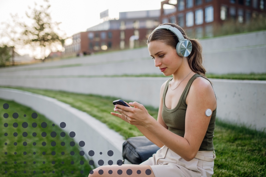 A young woman with long brown hair, wearing a green tank top and beige shorts, sits on outdoor concrete steps in a park-like setting. She wears stylish over-ear headphones and is focused on her smartphone, using the Cronometer app to track her continuous glucose monitor (CGM) data alongside her nutrition information. A CGM sensor is attached to her upper arm, helping her monitor blood glucose levels in real time.