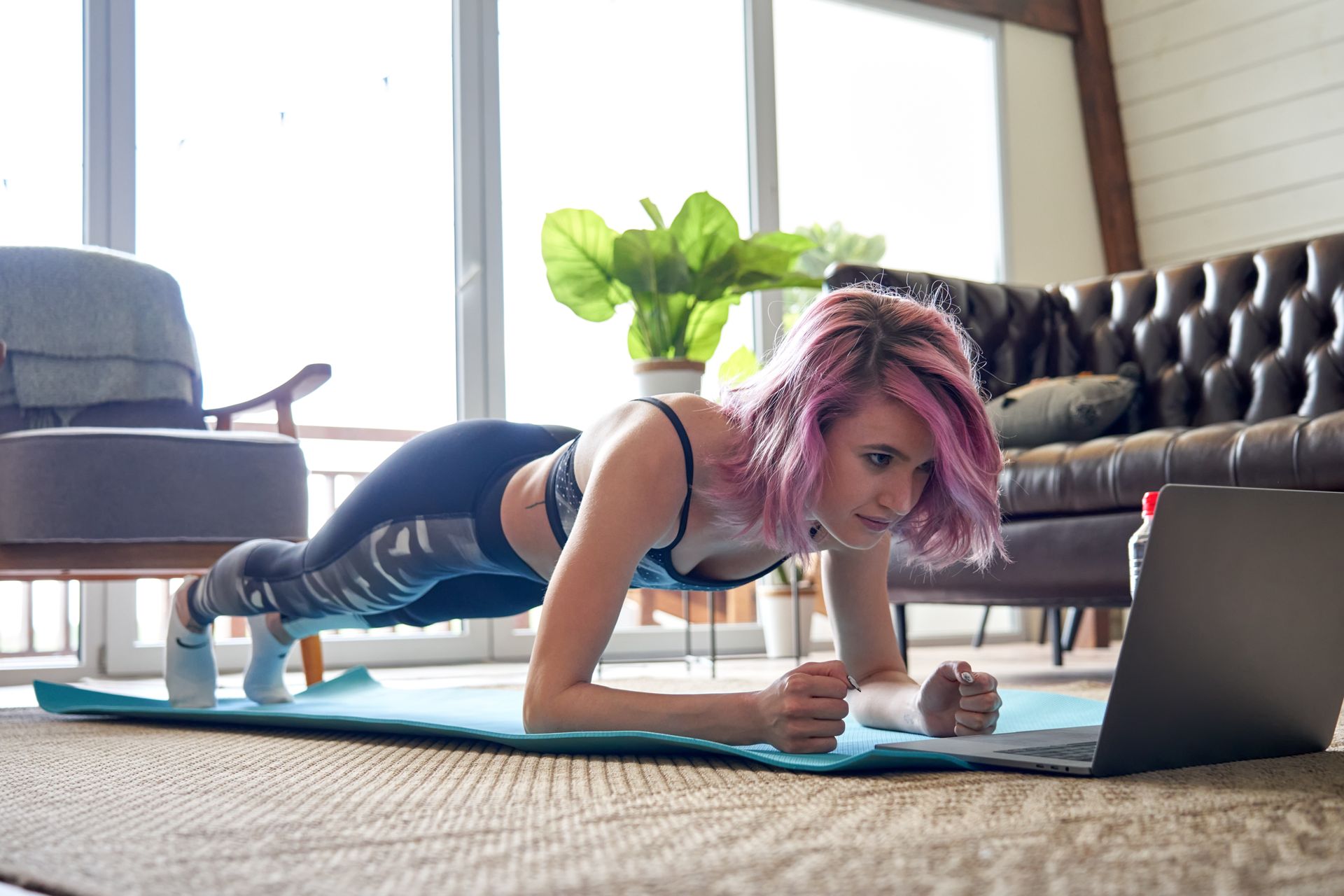 Sporty fit woman pink hair doing plank watching online class tutorial at home.