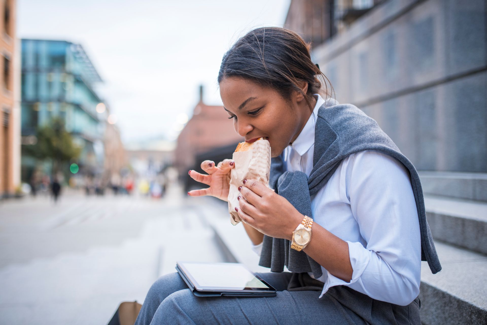 Businesswoman sitting on the stairs, eating sandwich and reading something at digital tablet.